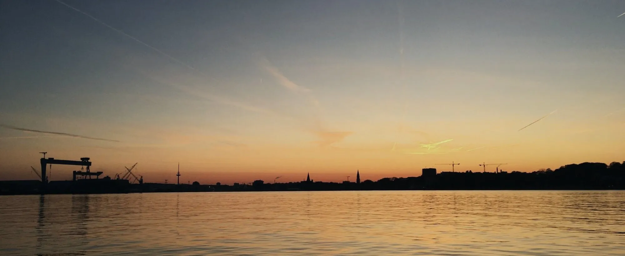 Die Kieler Förde am Abend, vom Wasser aus gesehen: die Silhouette der Stadt mit dem Werftkran am Horizont.